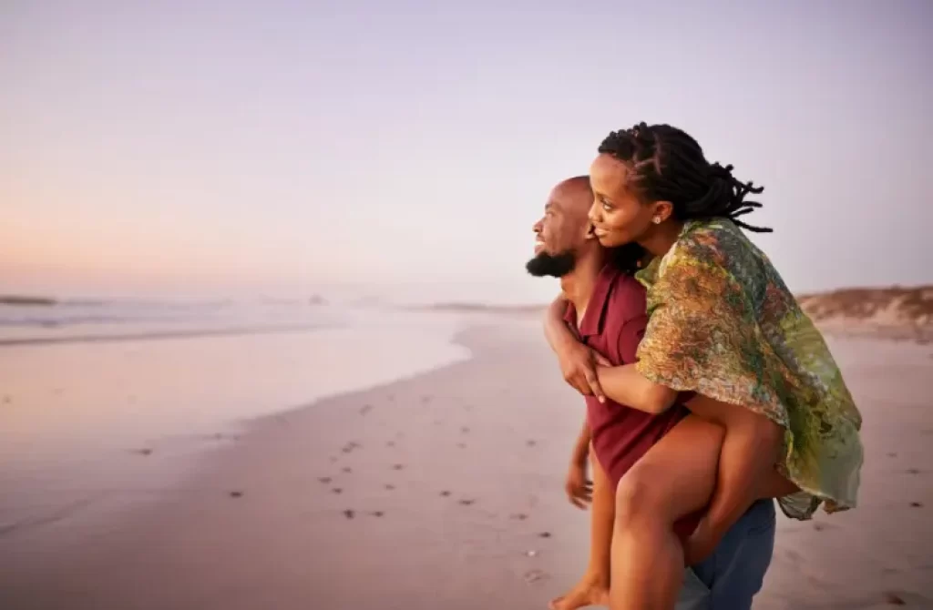 Pareja feliz disfrutando un momento en la playa al atardecer, hombre cargando a mujer en su espalda.